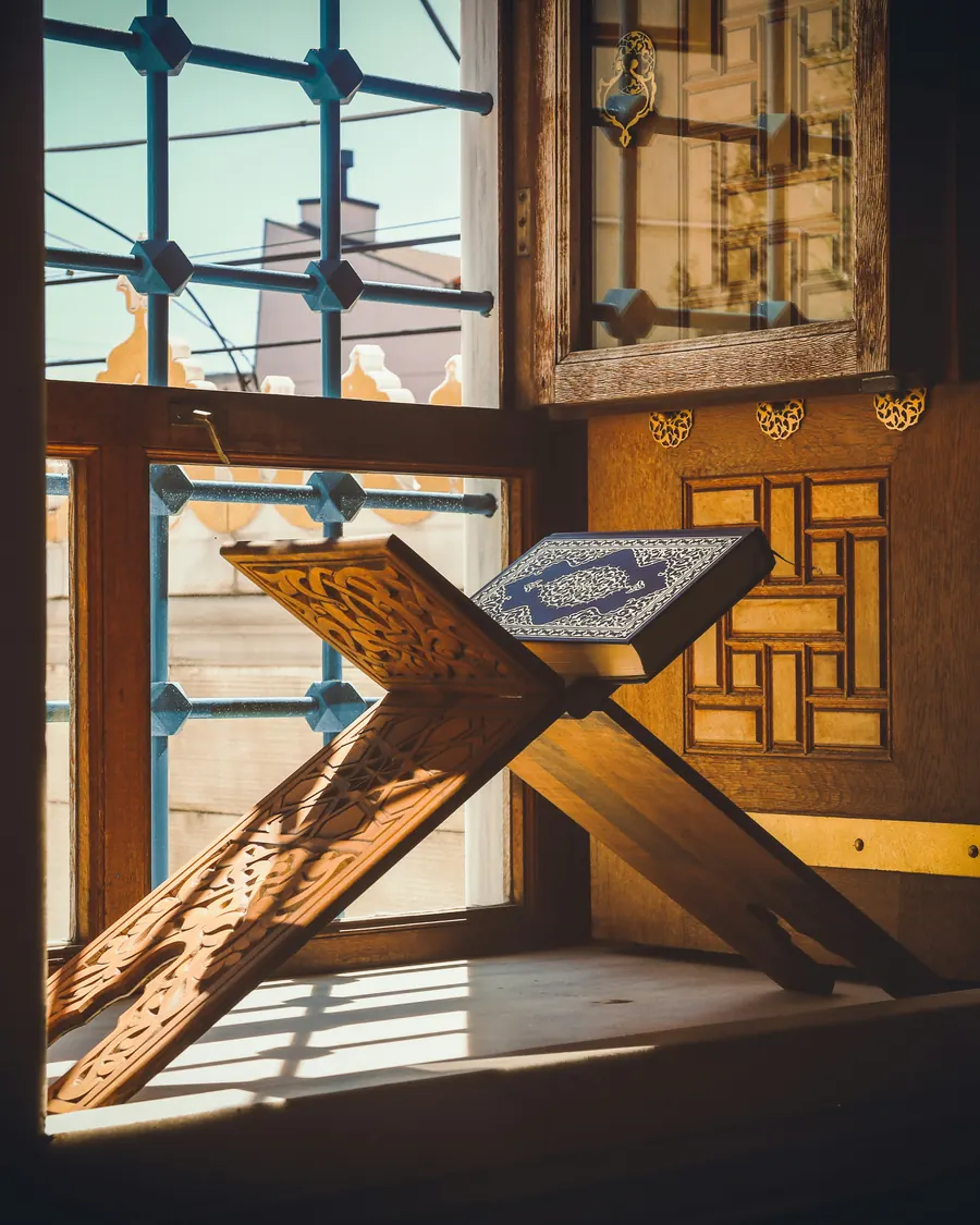 A Qur'an with a blue decorated cover resting on a carved wooden rehal on a sunlit windowsill beside a lattice window